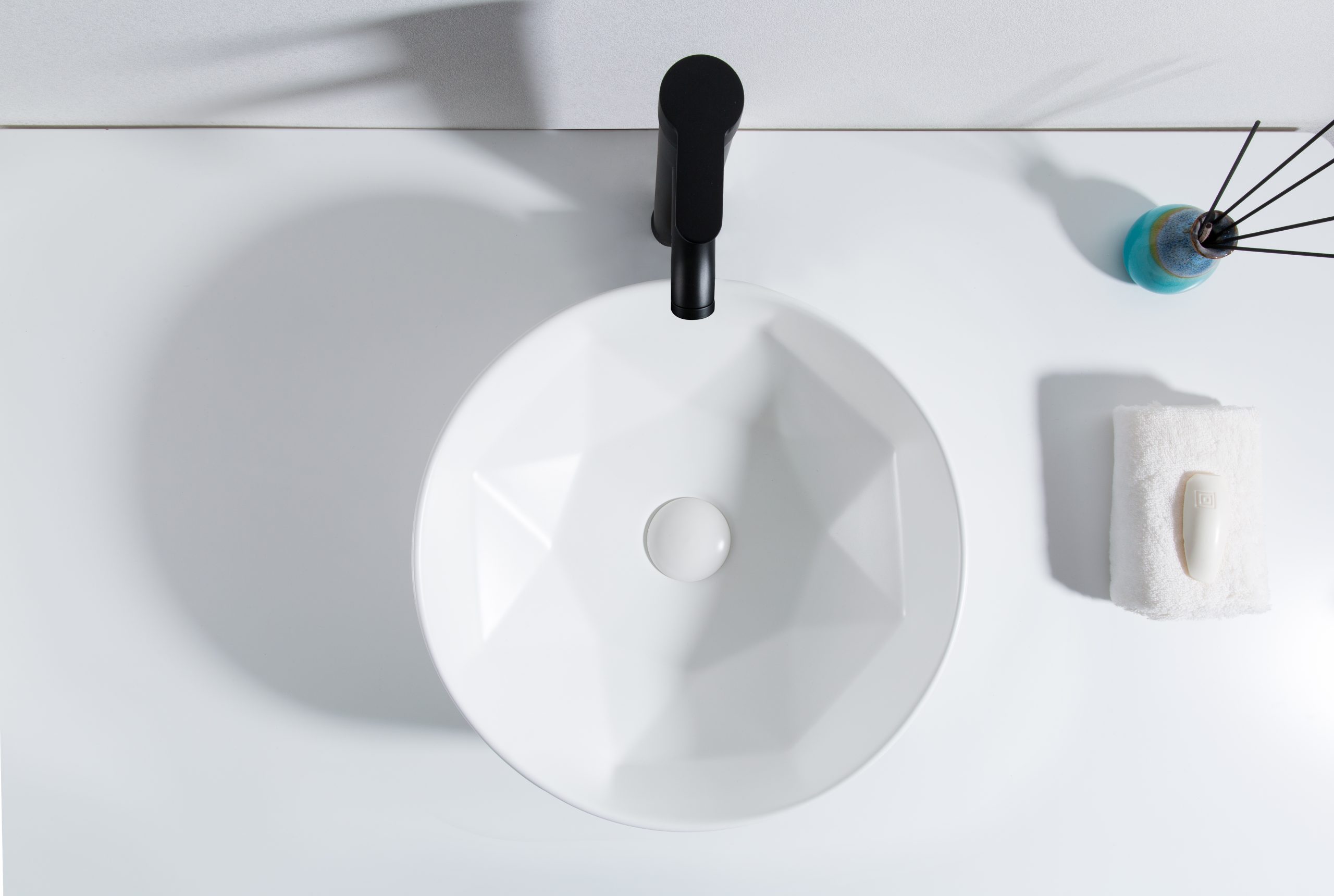 Bovenaanzicht van een ronde witte wastafel met geometrisch patroon, voorzien van een zwarte kraan, geplaatst op een witte ondergrond.